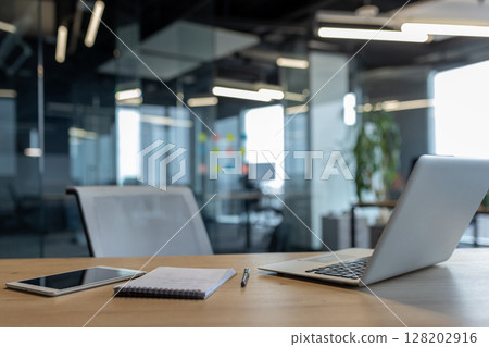 Contemporary workspace featuring a laptop, a smartphone, and a notebook on a wooden table. The background showcases a sophisticated office setting with glass panels and minimalistic design Contemporary workspace featuring a laptop, a smartphone, and a notebook on a wooden table. The background showcases a sophisticated office setting with glass panels and minimalistic design 128202916