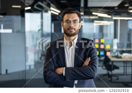 Portrait of serious confident businessman at workplace inside office. Man with crossed arms looking at camera, financier investor in business suit. Portrait of serious confident businessman at workplace inside office. Man with crossed arms looking at camera, financier investor in business suit. 128202918