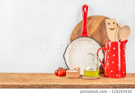 A cozy rustic kitchen scene with red and white cookware, wooden utensils, olive oil, and tomatoes ready for a homemade cooking experience 128203410