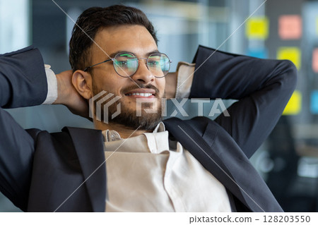 Confident young businessman wearing glasses and suit relaxing at office desk. Demonstrates relaxation, success, and contentment in professional setting. Ideal for business and lifestyle concepts. Confident young businessman wearing glasses and suit relaxing at office desk. Demonstrates relaxation, success, and contentment in professional setting. Ideal for business and lifestyle concepts. 128203550