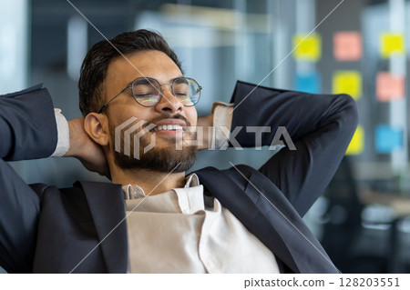 Successful businessman resting while sitting at his desk inside the office. Well done man with his hands behind his head with closed eyes smiling, satisfied with results of achieving the workplace. Successful businessman resting while sitting at his desk inside the office. Well done man with his hands behind his head with closed eyes smiling, satisfied with results of achieving the workplace. 128203551