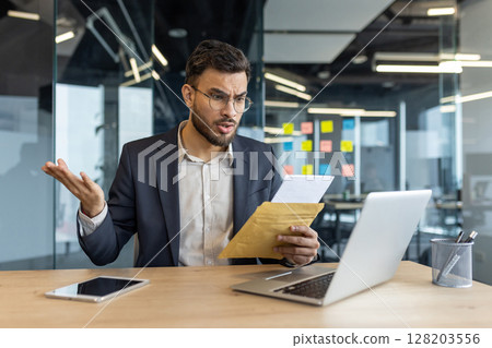 Mature businessman sits at a desk in office, reading a letter that appears to contain bad news. His expressive reaction displays concern or frustration. 128203556