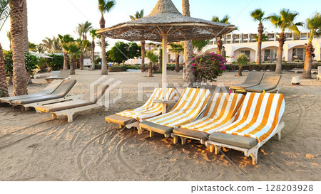 Beach resort in Egypt by the Red Sea. Sun loungers with orange and white stripes under a straw umbrella. Palm trees and sandy ground create a tropical atmosphere. Beach resort in Egypt by the Red Sea. Sun loungers with orange and white stripes under a straw umbrella. Palm trees and sandy ground create a tropical atmosphere. 128203928
