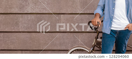 Closeup of unrecognizable african man standing near his bike, holding handlebar, posing over grey urban wall Closeup of unrecognizable african man standing near his bike, holding handlebar, posing over grey urban wall 128204060