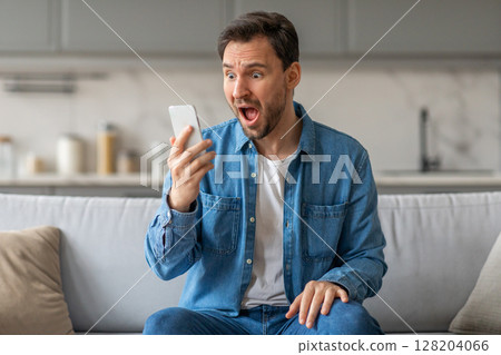A man in a denim shirt sits on a couch and stares at his smartphone with a look of shock and surprise on his face. His mouth is open, and his eyes are wide with disbelief 128204066