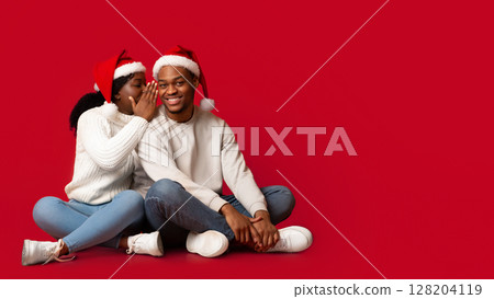 Romantic Black Woman Whispering To Boyfriend's Ear, Sharing Christmas Secret With Her Lover, Smiling African Couple Wearing Santa Hats, Having Fun Together While Sitting On Orange Background 128204119