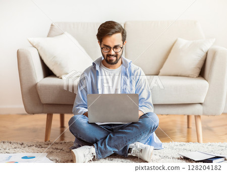 Remote Work Concept. Smiling Arab Freelancer Guy Using Laptop Computer At Home, Sitting On Floor In Living Room, Young Eastern Man Enjoying Working Remotely And Distant Job Opportunities, Copy Space 128204182