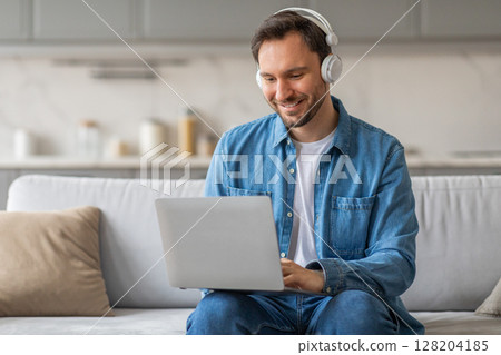 A man in a denim shirt sits on a couch in a living room. He is using a laptop computer and smiling. He is wearing headphones and is focused on his work. 128204185