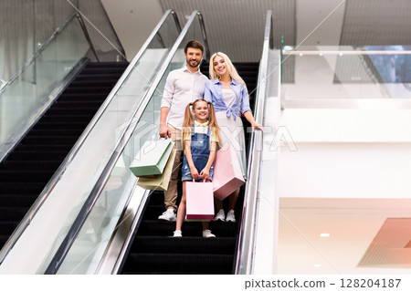 A happy family, consisting of a father, a mother, and a young girl, ride an escalator upward in a modern shopping mall. They are all smiling and holding shopping bags 128204187