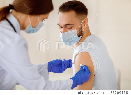 Female doctor making injection of coronavirus vaccine to young patient in medical mask at health centre. Millennial guy receiving vaccination against covid-19. Global immunization campaign 128204188