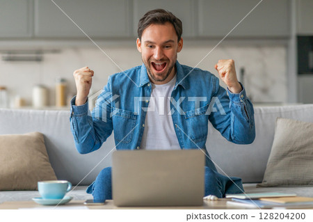 A man wearing a blue shirt and jeans is sitting on a sofa, looking at a laptop with a wide smile and both fists raised in the air. He seems to be celebrating a success or achievement 128204200