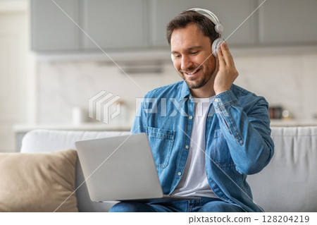 A man wearing headphones sits on a couch in a living room while smiling and using a laptop. He is wearing a blue denim shirt over a white t-shirt and blue jeans. A man wearing headphones sits on a couch in a living room while smiling and using a laptop. He is wearing a blue denim shirt over a white t-shirt and blue jeans. 128204219