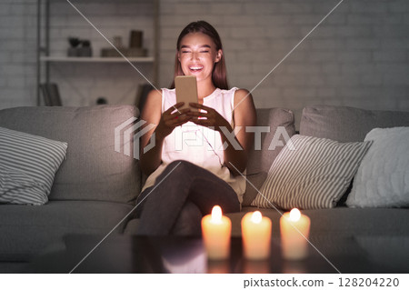 Happy Girl Texting On Cellphone Waiting For Boyfriend Sitting On Sofa In Dark Room Lit By Candles. Selective Focus Happy Girl Texting On Cellphone Waiting For Boyfriend Sitting On Sofa In Dark Room Lit By Candles. Selective Focus 128204220