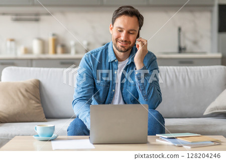 A man sits on a white couch in a living room and works on his laptop while speaking on a phone. He is wearing a blue jean shirt and smiling. On the table in front of him is a cup of coffee A man sits on a white couch in a living room and works on his laptop while speaking on a phone. He is wearing a blue jean shirt and smiling. On the table in front of him is a cup of coffee 128204246