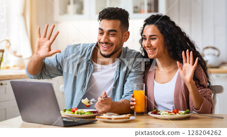 Cheerful arab couple making video call on laptop in kitchen, while having breakfast together, waving hands at camera, chatting with friends. Middle eastern spouses enjoying online communication Cheerful arab couple making video call on laptop in kitchen, while having breakfast together, waving hands at camera, chatting with friends. Middle eastern spouses enjoying online communication 128204262
