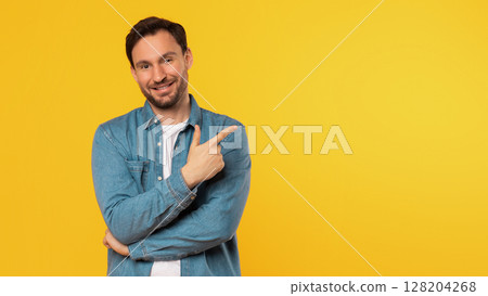 A man with short brown hair and a beard smiles at the camera while pointing at copy space. He is wearing a white t-shirt under a blue denim shirt, and he has his arms crossed. Background is yellow. 128204268