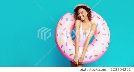 A young black woman with curly hair and a straw hat is posing with a donut pool float against a bright blue background. She is wearing a white swimsuit and smiling at the camera, copy space 128204291