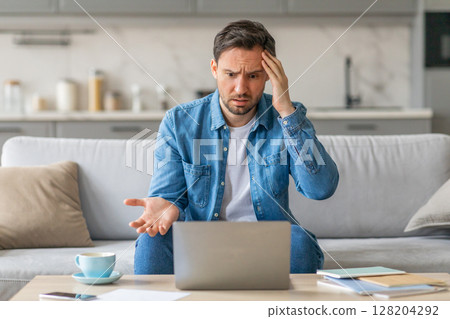 A man sits on a couch at home, looking frustrated and confused while using a laptop. He has one hand on his head and is gesturing with his other hand, as if he is trying to explain something. 128204292