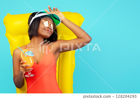 Pretty black lady in swimsuit lying on inflatable lilo and enjoying tropical cocktail over blue studio background. Beautiful African American woman relaxing on pool float, having summer chill 128204293
