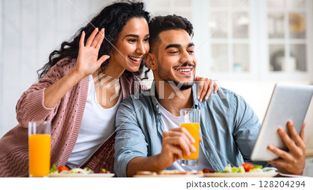 Modern muslim couple making video call on digital tablet in kitchen, while eating breakfast together, smiling middle eastern couple waving hands at camera, enjoying online communication, free space 128204294