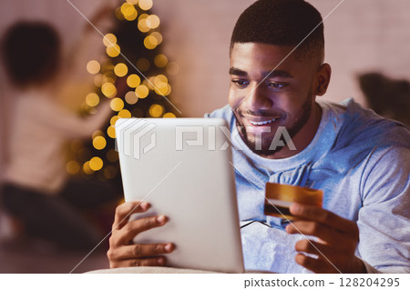 Afro man ordering xmas gifts on tablet, woman decorating Christmas tree on background 128204295