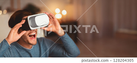 Excited Millennial Guy Using Virtual Reality Glasses Playing Videogame At Home In The Evening. Selective Focus Excited Millennial Guy Using Virtual Reality Glasses Playing Videogame At Home In The Evening. Selective Focus 128204305