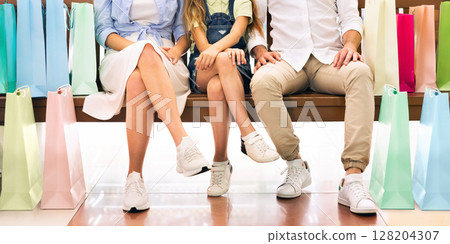 A family of three, a man, a woman, and a child, sit on a bench with many colorful shopping bags at their feet, likely after a shopping spree in a retail store, cropped, panorama A family of three, a man, a woman, and a child, sit on a bench with many colorful shopping bags at their feet, likely after a shopping spree in a retail store, cropped, panorama 128204307