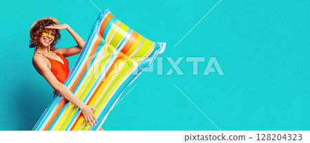 Beautiful African American woman in swimsuit, sunglasses and summer hat holding inflatable raft, isolated on blue background, panorama with copy space 128204323
