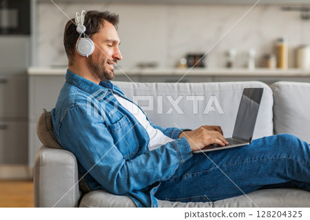 A man in a denim shirt sits on a gray couch with his legs stretched out. He is wearing white headphones and looking down at his laptop while typing. A man in a denim shirt sits on a gray couch with his legs stretched out. He is wearing white headphones and looking down at his laptop while typing. 128204325