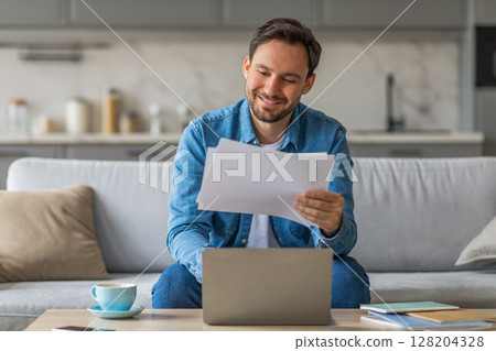 A man is sitting on a sofa, smiling as he reviews some documents while using a laptop computer. He is wearing a blue denim shirt, and the sofa is in a home living room. 128204328