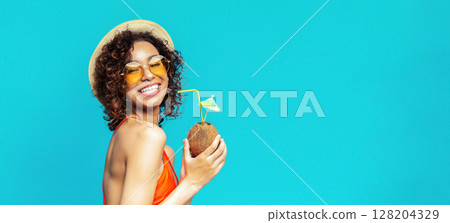 African American young woman with curly hair is smiling while holding a coconut with a straw in her hand. She is wearing a straw hat and sunglasses. The background is a bright blue. 128204329