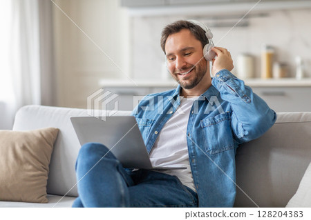 A man sits on a white couch in his living room, wearing a denim shirt, jeans, and headphones. He smiles as he looks at his laptop and adjusts his headphones with one hand. A man sits on a white couch in his living room, wearing a denim shirt, jeans, and headphones. He smiles as he looks at his laptop and adjusts his headphones with one hand. 128204383