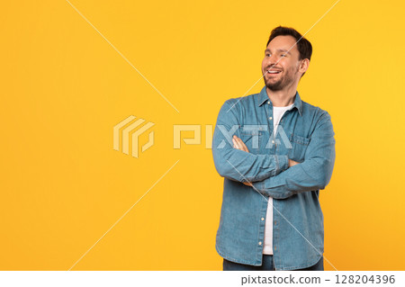A man with a beard and short brown hair wears a blue denim shirt with a white undershirt. He stands in front of a yellow background, smiling, with his arms crossed. He is looking at copy space 128204396