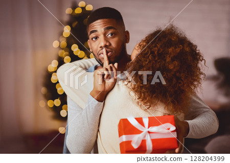 Shh. Afro man giving Christmas present to woman, embracing her and making silence sign Shh. Afro man giving Christmas present to woman, embracing her and making silence sign 128204399
