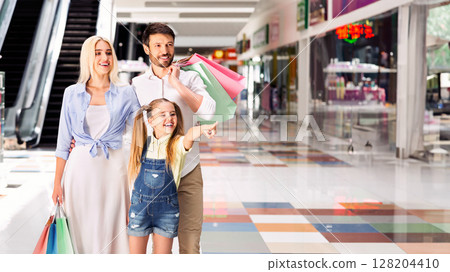 A young family, consisting of a mother, father, and daughter, is walking through a shopping mall. They are holding colorful shopping bags and appear to be happy, copy space A young family, consisting of a mother, father, and daughter, is walking through a shopping mall. They are holding colorful shopping bags and appear to be happy, copy space 128204410