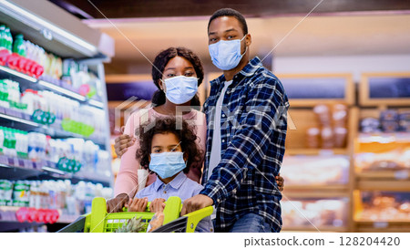Portrait of young black family with little child wearing face masks while shopping for groceries at supermarket during covid outbreak. Millennial parents and their daughter buying food at mall 128204420