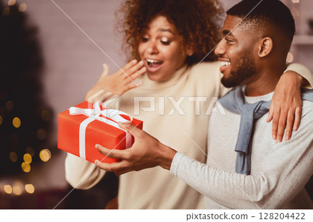Surprised black woman getting Christmas present from husband, celebrating holidays together at home 128204422