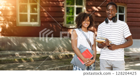 Smiling black guy and girl university students standing outdoors near campus with books and workbooks in hands and posing to camera, free space 128204425