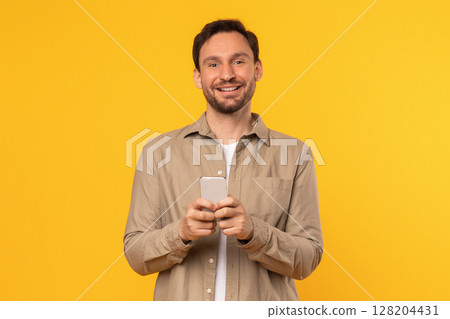 A man wearing a beige shirt stands against a bright yellow background, smiling as he holds a smartphone in his hands. 128204431