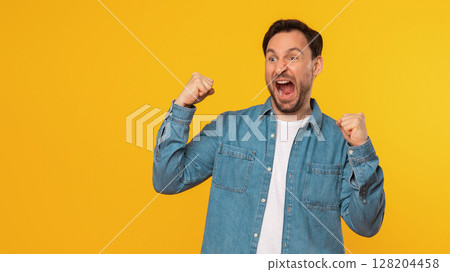 A man wearing a blue denim shirt with a white t-shirt underneath celebrates his success, with a big grin and both fists raised in the air. He stands against a bright yellow background, copy space 128204458