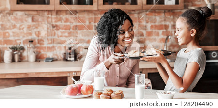 Happy little african girl showing self made croissants to her cheerful mom, kitchen interior Happy little african girl showing self made croissants to her cheerful mom, kitchen interior 128204490