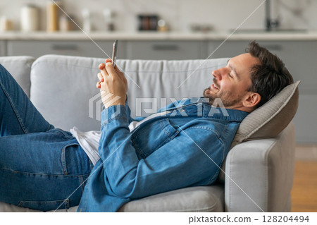 A man lies on a grey couch, propped up on a pillow, with his legs outstretched and his arms resting on the couch. He is using his smartphone with a smile on his face. A man lies on a grey couch, propped up on a pillow, with his legs outstretched and his arms resting on the couch. He is using his smartphone with a smile on his face. 128204494