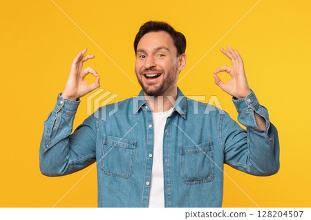 A smiling man wearing a blue denim shirt is standing against a bright yellow background. He is making the okay gesture with both hands. 128204507
