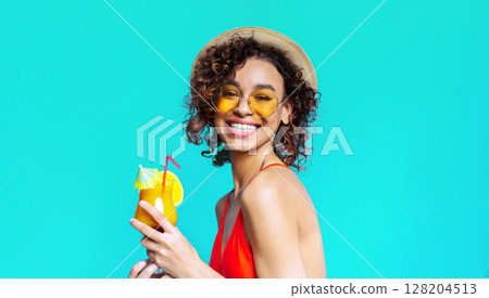 African American woman with curly brown hair wears a straw hat and sunglasses, while smiling and holding a cocktail. She is wearing a red swimsuit and is standing in front of a turquoise background. 128204513