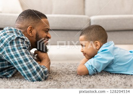 Happy African Father And Son Looking At One Another Lying On Floor At Home. Dad And Kid Bonding. Child And Daddy's Relationship And Trust Concept. Side View Portrait 128204534