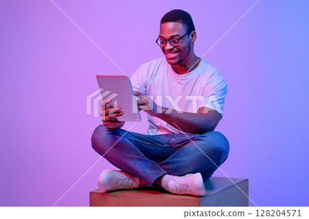 Young black freelancer guy in glasses sitting with digital tablet under luminous neon light, african american man blogger working online, browsing internet and enjoying modern techologies, copy space 128204571