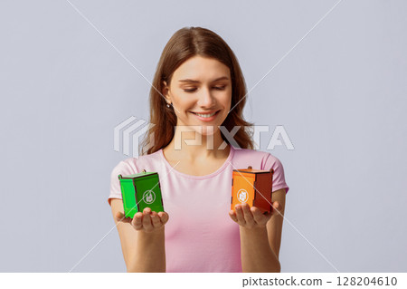 A woman displays two colorful recycling bins, smiling while encouraging eco-friendly practices. Her casual clothing and the neutral background set a positive tone for sustainability awareness. 128204610