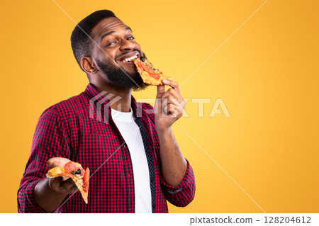 Black Guy Enjoying Eating Tasty Pizza Standing Over Yellow Studio Background. Junk Food Overeating. Fastfood Eater Biting Slice Of Unhealthy Pizza. Nutrition And Binge Eating Habit Concept 128204612