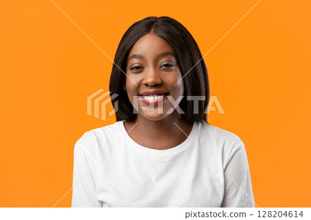 Closeup portrait of cute cheerful black young lady in white t-shirt posing on orange studio background, copy space. Beautiful african american woman with shining fair hair smiling at camera, flirting 128204614