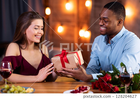 Caring African American Guy Surprising His Girlfriend With Wrapped Gift Box On Romantic Date In Restaurant. Loving Multiracial Couple Dining Together In Elegant Atmosphere, Celebrating Valentine's Day 128204627